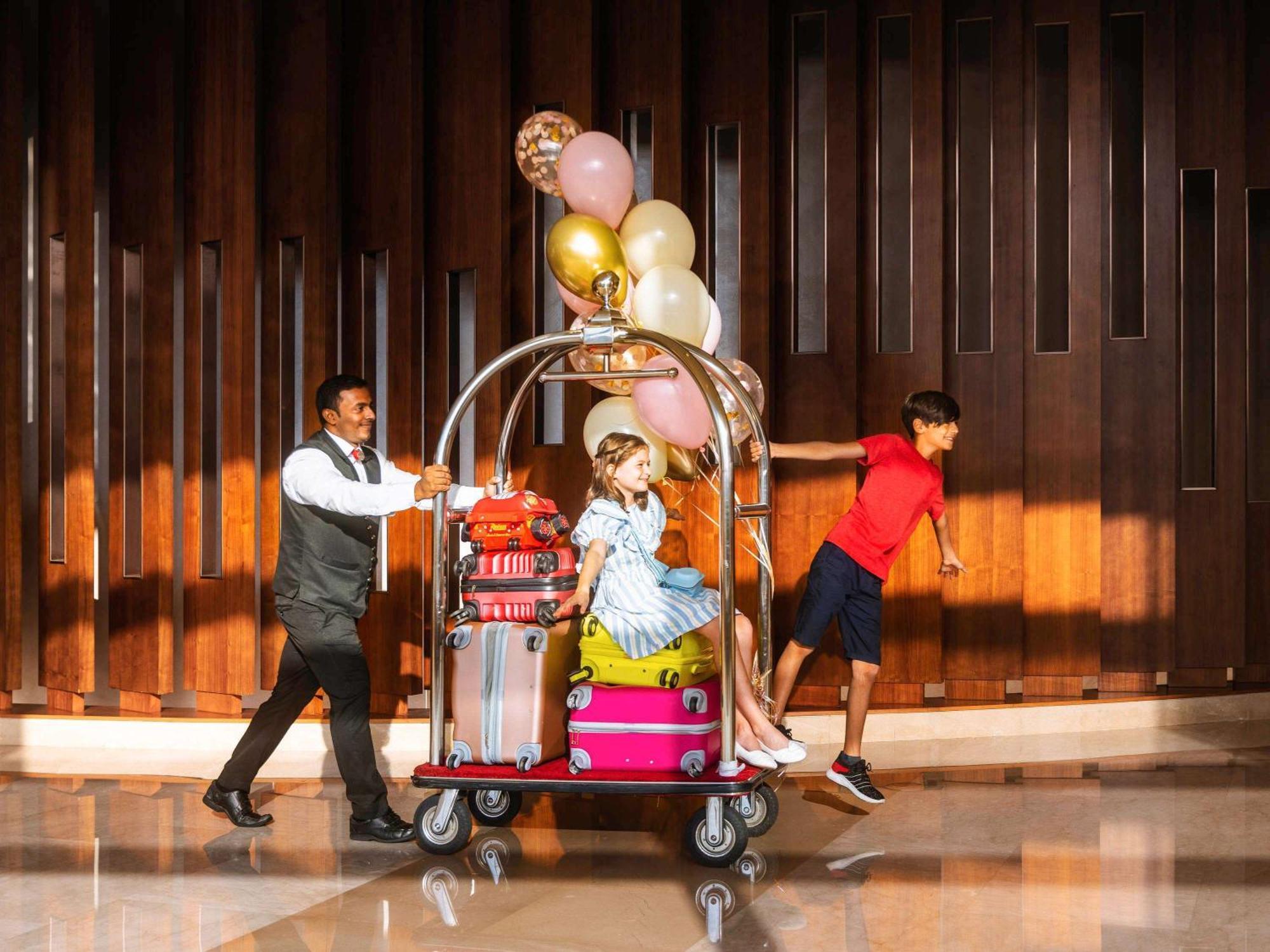 Swissôtel Al Ghurair Dubai Bagian luar foto The photo depicts a hotel scene where a bellhop is pushing a luggage cart. The cart is piled high with various colorful suitcases, and it is topped with balloons in shades of pink, gold, and white. On the cart sits a girl dressed in a light blue dres